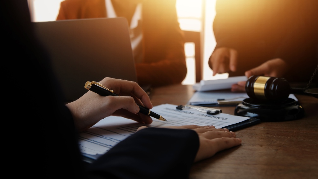 Person signing contract papers with a pen, wooden gavel and another individual in background.