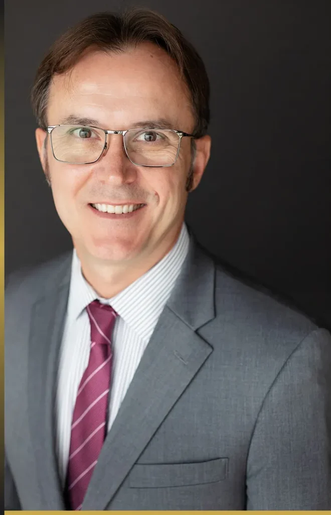 Professional middle-aged man wearing glasses, gray suit, striped shirt, and maroon tie, smiling against black background.