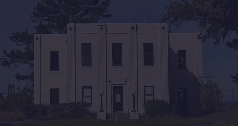 Two-story beige building with symmetrical windows and central entrance, surrounded by trees.